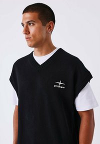 Chaleco de punto negro con un diseño de cuello en V, con acentos de canalé y un logotipo plateado "PXP" en el lado izquierdo, llevado sobre una camiseta blanca.