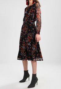 Černé šaty s dlouhými průsvitnými rukávy, elastickým pasem a barevným květinovým vzorem. Kombinované s černými kotníkovými botami.