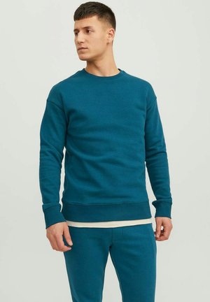 Felpa color teal con girocollo, polsini e vita a costine. Materiale in cotone con una texture liscia. Indossata con pantaloni abbinati.
