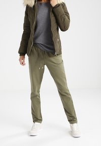 Chaqueta de plumas verde oliva con capucha de pelo sintético, camiseta gris y joggers sueltos en verde oliva combinados con zapatillas blancas.