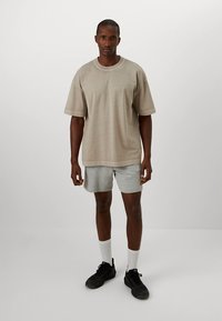 Bézs rövid ujjú póló, szürke sport rövidnadrággal párosítva. A póló kerek nyakkivágású, a rövidnadrág enyhén texturált. Fekete sportcipő és fehér zokni teszik teljessé az öltözéket.