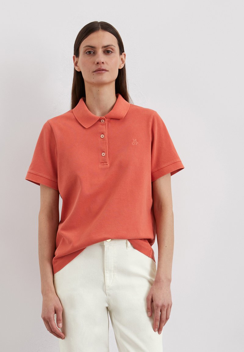 Koraalkleurig poloshirt met een kraag, drie knopen en een klein geborduurd logo. Gepaarde met witte broek, met een zachte textuur.