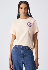 Perzikkleurig katoen T-shirt met korte mouwen, voorzien van een paarse grafische logo op de linkerborst. Gecombineerd met hooggetailleerde blauwe spijkerbroeken.
