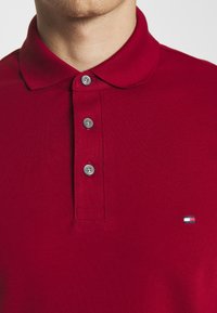 Uomo che indossa una polo rossa con tre bottoni e un piccolo logo rettangolare sul petto su sfondo uniforme.