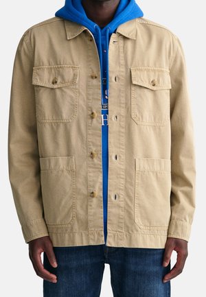 Uomo che indossa una giacca beige con quattro tasche abbottonata sopra una felpa con cappuccio blu e jeans blu, in piedi contro uno sfondo chiaro.