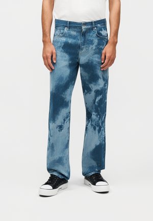 Homme portant un jean tie-dye bleu et des baskets noires, debout contre un fond blanc uni.