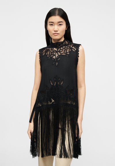 Γυναίκα που φοράει αμάνικο μαύρο δαντελωτό τοπ με λουλουδένια σχέδια και μακριές φούντες πάνω σε ανοιχτόχρωμο παντελόνι, στέκεται μπροστά σε απλό φόντο.