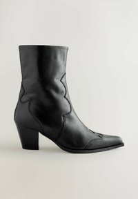 Bota western de cuero negro hasta la mitad de la pantorrilla con punta afilada, costura decorativa y tacón cuadrado sobre un fondo gris claro.