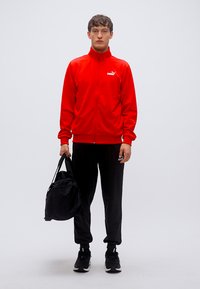 Κόκκινο μπουφάν με φερμουάρ και ψηλό γιακά, μαύρο αθλητικό παντελόνι και μαύρη τσάντα. Το μοντέλο φοράει μαύρα αθλητικά παπούτσια και στέκεται σε ουδέτερο φόντο.