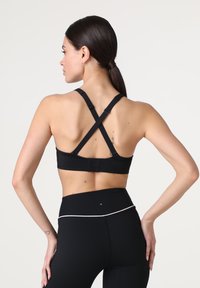 Top sportivo nero con spalline incrociate, texture liscia, chiusura posteriore regolabile e design minimalista, abbinato a leggings neri a vita alta.