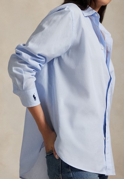 Cămașă albastru deschis cu nasturi, realizată din bumbac. Are o croială lejeră, mâneci suflecate și un mic logo pe manșetă. Tivul este mai lung în partea din spate.