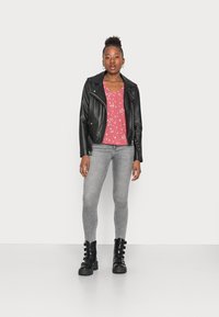 Jaqueta de motociclista de couro preto, blusa floral rosa, jeans skinny cinza e botas de tornozelo pretas com fivelas. Modelo está posicionada contra um fundo branco.