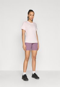 Ανοιχτό ροζ μπλουζάκι με λευκό λογότυπο, συνδυασμένο με μοβ ποδηλατικό σορτς. Το μοντέλο φοράει μαύρα παπούτσια και λευκές κάλτσες αστραγάλου, στέκεται σε απλό φόντο.