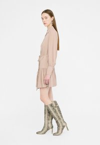Abito camicia beige con maniche lunghe, vita cintata e orlo arricciato, abbinato a stivali metallici in stampa serpente a texture al ginocchio.