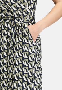 Granatowa sukienka w liściasty wzór w białych i oliwkowych odcieniach; charakteryzuje się wiązanym pasem i boczną kieszenią dla funkcjonalności.