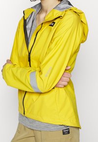 Keltainen vedenpitävä takki, jossa harmaa hupun vuori, edessä vetoketju ja heijastavat hihansuun yksityiskohdat. Kevyt materiaali; rento istuvuus.