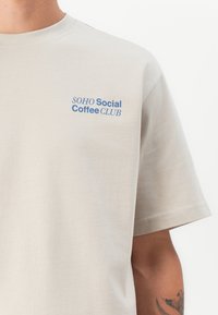 Beige Baumwoll-T-Shirt mit einem blauen Drucklogo auf der linken oberen Seite, das "SOHO Social Coffee CLUB" liest. Kurze Ärmel, runder Ausschnitt.