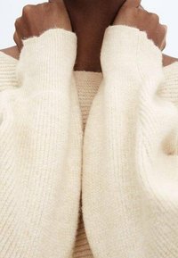 Un pull crème présente un design tricoté texturé avec des poignets larges en côtes et une coupe décontractée. Le tissu semble doux et confortable.