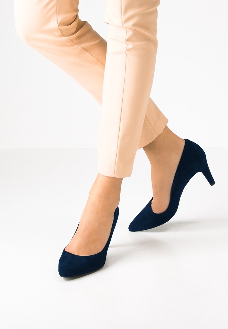 Femme portant un pantalon beige et des escarpins en suède bleu marine à talons aiguilles, se tenant sur un sol blanc.
