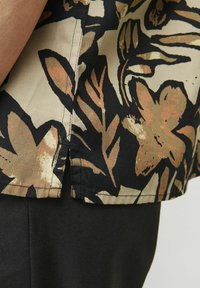 Chemise à manches courtes en beige avec un motif floral noir. Dispose d'un ourlet droit, de surpiqûres contrastées et d'un matériau en coton léger.
