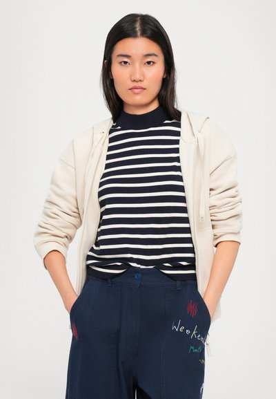 Young woman with straight dark hair wears navy striped shirt, cream zip-up hoodie, and navy pants with colorful embroidered text and designs.