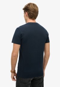Mand med lyst brunt hår iført en navy blå t-shirt med korte ærmer og sorte jeans, stående med ryggen til kameraet.