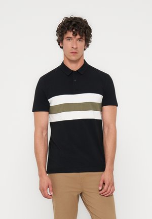 Mann mit lockigem dunklem Haar, der ein schwarzes Polohemd mit weißen und olivgrünen horizontalen Streifen trägt, kombiniert mit hellbrauner Hose, stehend vor weißem Hintergrund.