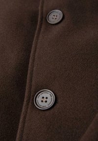 Lähikuva ruskeasta kankaasta, jossa on kaksi nelireikäistä nappia, joista toisessa on reunassaan teksti "OMBRE QUINTESSENCE".