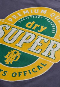 Grijze T-shirt met een grote, cirkelvormige afbeelding in geel en groen. De tekst omvat "Super," "droog," en "Premium Kwaliteit" in vette letters.