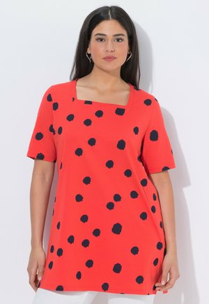 Frau trägt ein rotes Tunika mit schwarzen, unregelmäßigen Punkten, kurzen Ärmeln und quadratischem Ausschnitt, posiert vor einem schlichten, hellen Hintergrund.