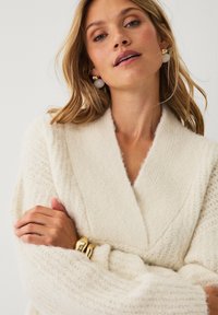 Cremefarvet, struktureret strikket cardigan med bred sjalskrave. Modellen bærer guldarmbånd og store, runde øreringe.