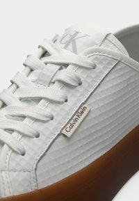 Baskets texturées blanches Calvin Klein avec lacets blancs, étiquette de marque sur le côté et semelle en caoutchouc marron en vue rapprochée.