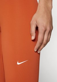 Pomarańczowe legginsy z gładkiego materiału, z białym logo Nike, zbliżenie przedstawiające dłoń opartą na udzie.