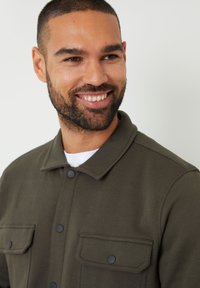 Chaqueta de color verde oliva con cuello y tejido texturizado, que presenta dos bolsillos en el pecho y cierres de botones negros, superpuesta sobre una camisa blanca.