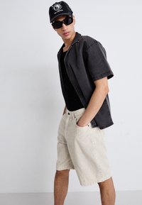 Sort kortærmet skjorte over en sort tanktop, parret med lysebeige fløjlsshorts. Modellen bærer sorte solbriller og en sort kasket.