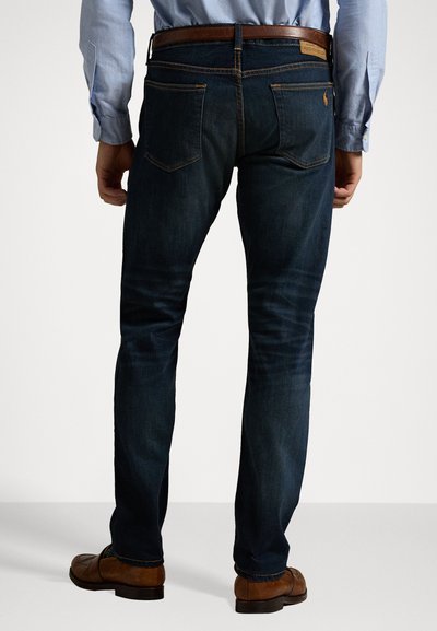 Jeans de mezclilla azul oscuro con un ajuste slim, que presentan bolsillos traseros, costuras visibles y un cinturón de cuero marrón con una hebilla rectangular.