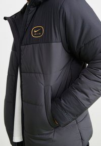 Svart isolerad jacka med dragkedja, guld Nike AIR-logotyp, slät textur, sidoficka och "THERM"-bokstäver.