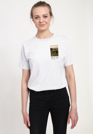 Giovane donna con capelli legati indossa una maglietta bianca con illustrazione di una corsa di cavalli e testo "Edgar Degas", con pantaloni neri su sfondo semplice.