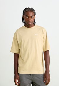 Beige katoenen t-shirt met korte mouwen en een ronde halslijn, met een wit Puma-logo op de borst. G gedragen met grijze jeans.
