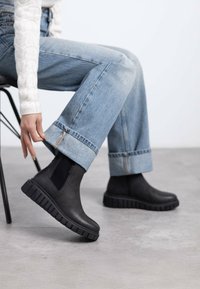 Stivali neri alla caviglia con suola in gomma spessa, pannelli laterali elastici e una texture liscia, abbinati a jeans azzurri a risvolto.