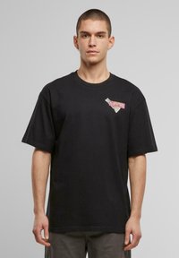 Black cotton t-shirt with a relaxed fit, featuring a graphic patch in pink and beige on the left chest area. Short sleeves.