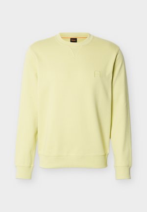 Lys gul crewneck-genser med ribbestrikkede mansjetter og nederkant, liten preget logo på venstre bryst, og svart halsmerke med oransje kant.