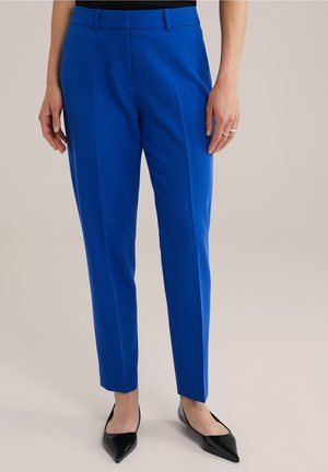 Femme portant un pantalon bleu vif sur mesure à la cheville et des ballerines noires pointues, debout devant un fond neutre.