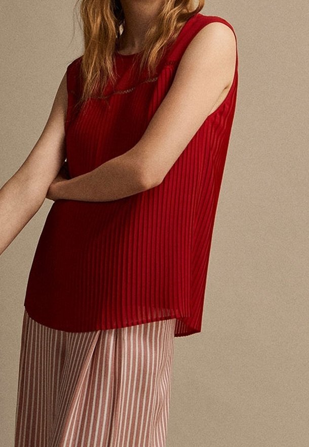 Haut rouge sans manches à plis porté avec un pantalon ample beige et blanc à rayures verticales sur une personne debout devant un fond uni.