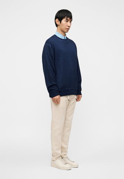 Φούτερ σε ναυτικό μπλε με στρογγυλή λαιμόκοψη, φορεμένο πάνω από ένα ανοιχτό μπλε πουκάμισο με γιακά. Συνδυασμένο με μπεζ παντελόνι και κρεμ παπούτσια.