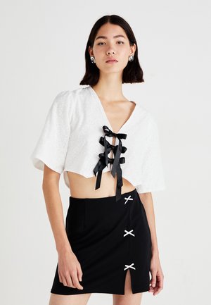 Hvit cropped bluse med korte ermer, hekledetaljer og svarte satengsnører. Svart miniskjørt med sløyfefordelinger og en側splitt.