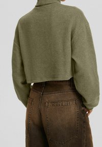 Oříznutý olivově zelený pletený svetr s vysokým límcem a žebrovanou strukturou, doplněný hnědými džínami s vysokým pasem a zadními kapsami.