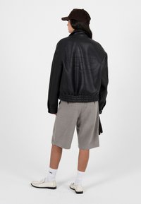 Osoba v černé kožené bundě, šedých kraťasech po kolena, bílých ponožkách, krémových loaferech a tmavě hnědé čepici, otočená zády ke kameře.