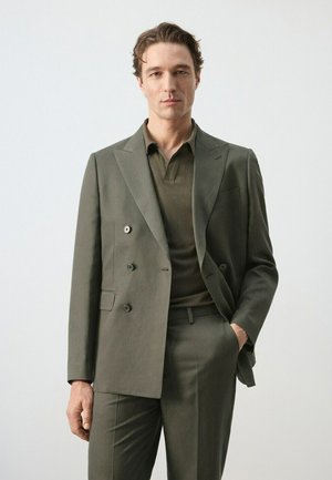 Uomo che indossa un abito doppiopetto verde oliva e una polo abbinata, in piedi con una mano in tasca su sfondo semplice.