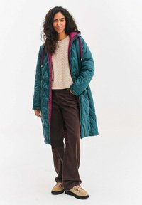 Dlouhý tyrkysový prošívaný kabát s vínovou podšívkou, doplněný béžovým svetr s copánkovým vzorem a hnědými kalhotami z manšestru. Outfit doplňují béžové turistické boty.